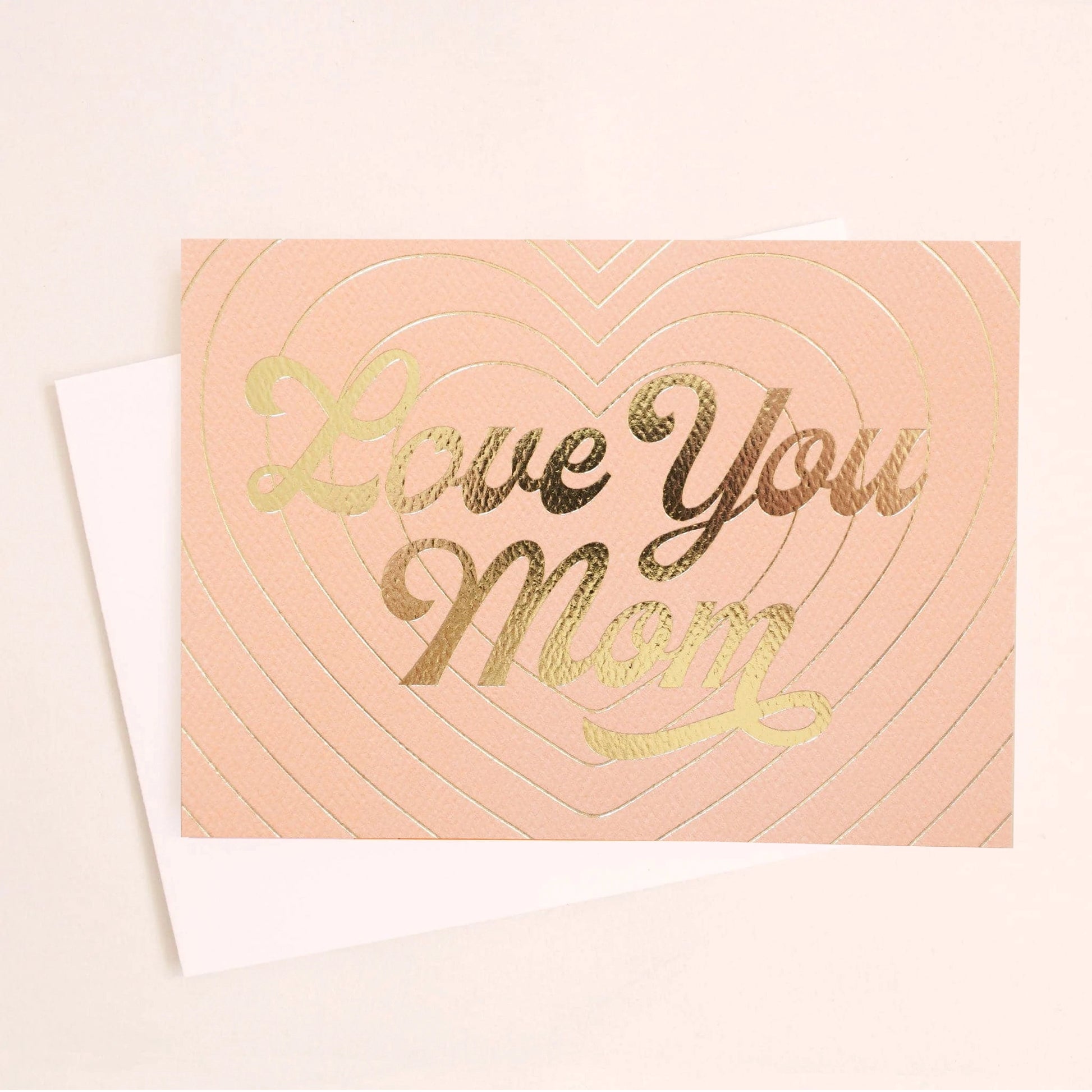 Peach card that reads 'love you mom' in gold foil. The card is filled with layers of a beaming gold foil heart design behind and is accompanied by with a white envelope. 