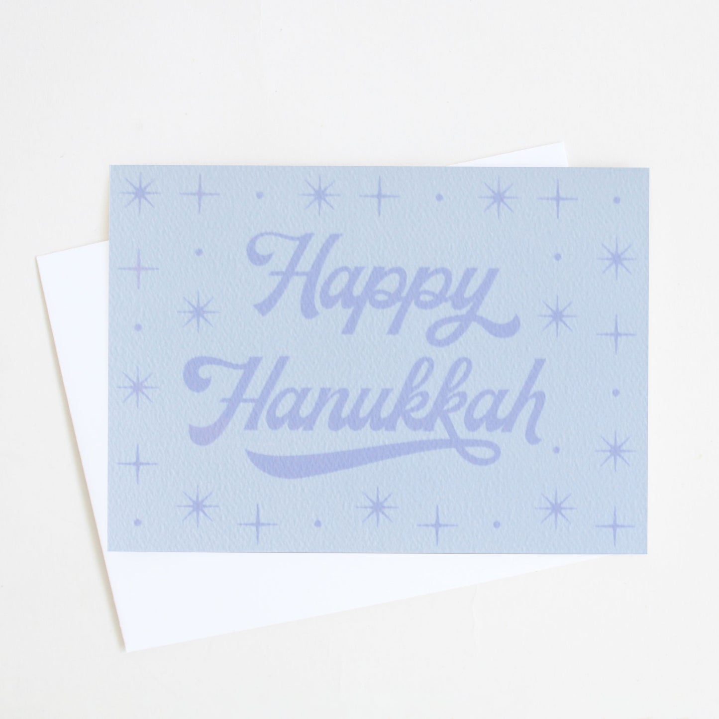 A light blue holiday card that reads, "Happy Hanukkah" along with various star designs and accompanied by a white envelope.