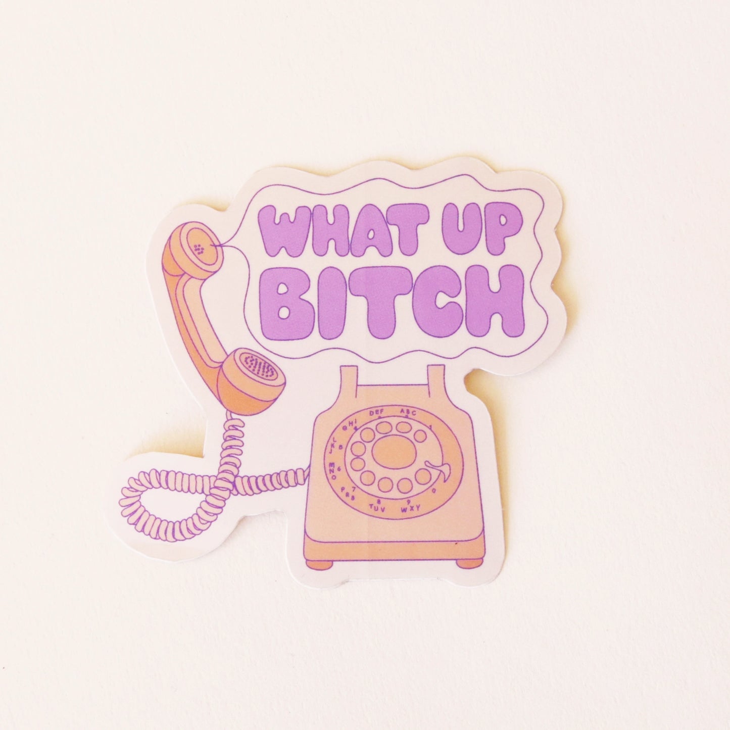On a cream background is a vinyl sticker of an old school pink phone with a speech bubble coming out of the phone that reads, "What Up Bitch" in purple letters.